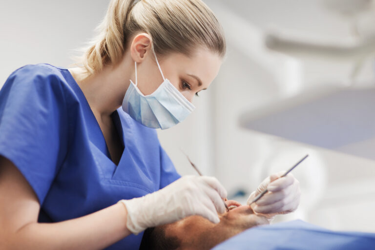 people, medicine, stomatology and health care concept - female dentist with dental mirror and probe checking up male patient teeth at dental clinic office